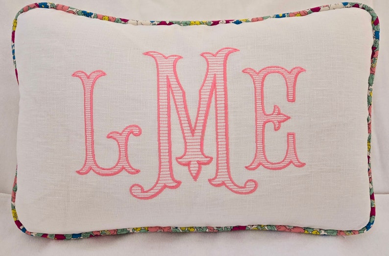 Może przedstawiać: Biała poduszka z haftowanym r&oacute;żowym monogramem "LME" na białym materiale lnianym. Poduszka ma ozdobny brzeg z kwiatowym wzorem.