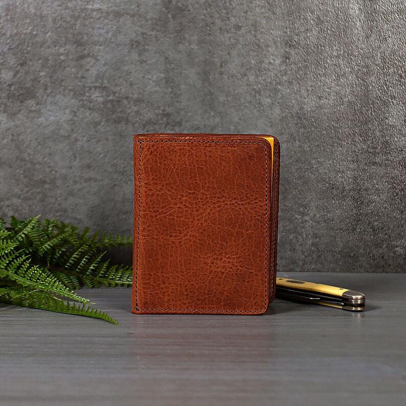 May include: A brown leather journal with a textured surface. The journal is closed, with a yellow edge visible. A small pocket knife rests beside the journal on a gray surface. Fern fronds are in the background.