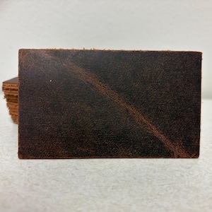 May include: Close-up of a rectangular piece of distressed brown leather. The leather has a textured surface with darker and lighter brown variations. A stack of similar leather pieces is visible in the background. The leather appears to be for crafting.