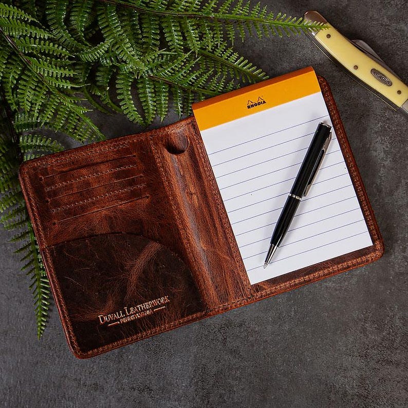 May include: Open brown leather notebook cover with a notepad and a black pen. The notepad has an orange top and white lined paper. A small pocket knife is in the background. The leather cover has the text "DUVALL LEATHERWORK" on the bottom.