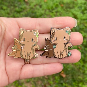 May include: Two enamel pins depicting a cartoon cat beside a tree stump. One pin is gold-toned with a light brown cat and stump, the other is silver-toned with a dark brown stump and cat. Both have floral details.