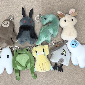 May include: A collection of nine plush keychains featuring various characters including a brown sasquatch, a dark grey moth-like creature, a green dinosaur, a cream-coloured rabbit, a white ghost, a green frog, a yellow pig, a house, and a white yeti.