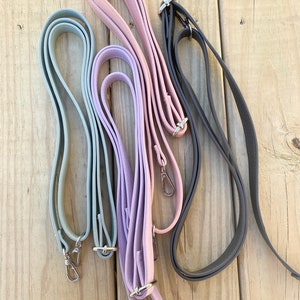 May include: An assortment of five colorful purse straps in shades of gray, pink, and purple. Each strap is made of a smooth, flexible material and features silver-tone metal hardware, including clasps and rings for attachment.