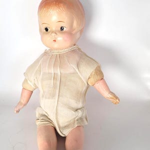 May include: Vintage doll with a painted face, short red hair, and a cream-colored outfit. The doll has a detailed face with painted eyes and a small mouth. It wears white shoes with black trim. A collectible item.