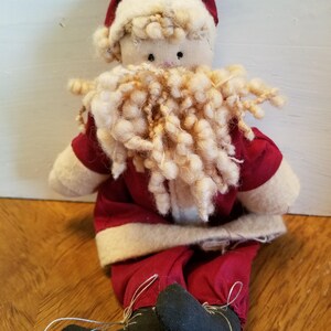 May include: A handmade rag doll Santa Claus with a red suit, white trim, and a fluffy white beard. The doll is sitting with its legs crossed and wearing black shoes.