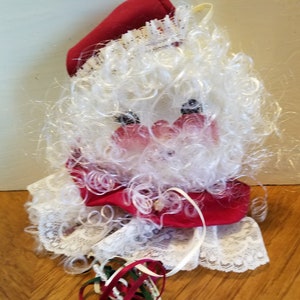 Peut inclure: Une décoration de Noël représentant un Père Noël blanc et rouge avec une bordure en dentelle blanche et une longue chaîne de perles et de cloches.