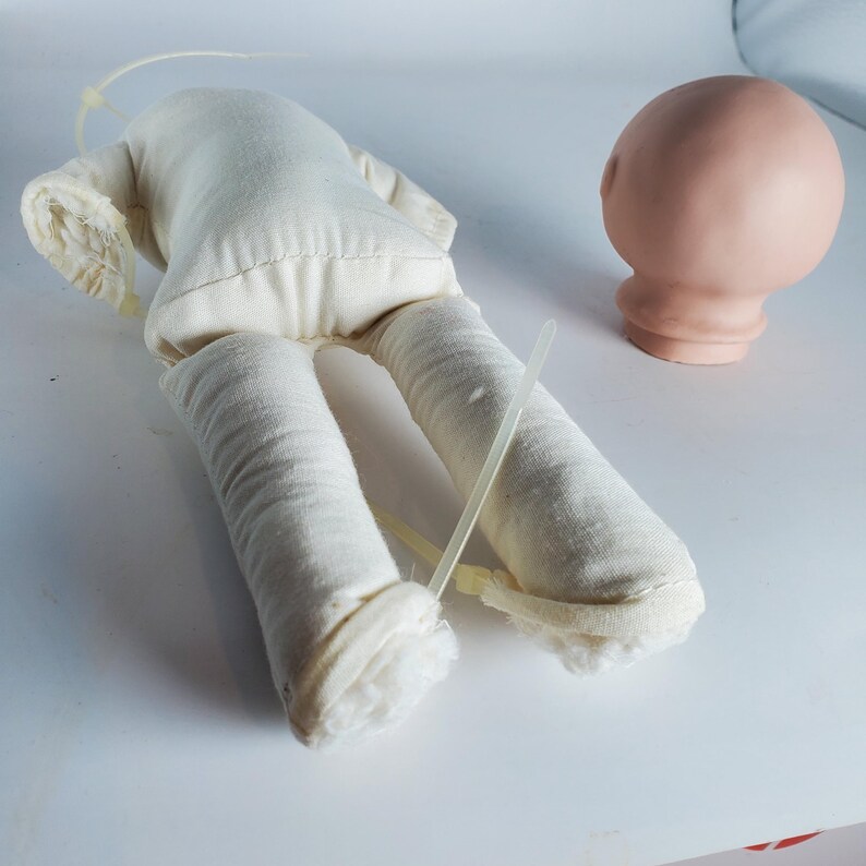 May include: A doll-making kit featuring a cream-colored fabric doll body with attached arms and legs, and a separate, unpainted, peach-colored doll head. The body is stitched together, and the limbs are stuffed. Plastic ties are attached.