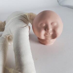 May include: A vintage doll head with a painted face and a cloth body. The doll head is a light pink color with closed eyes and a slight smile. The cloth body is white.