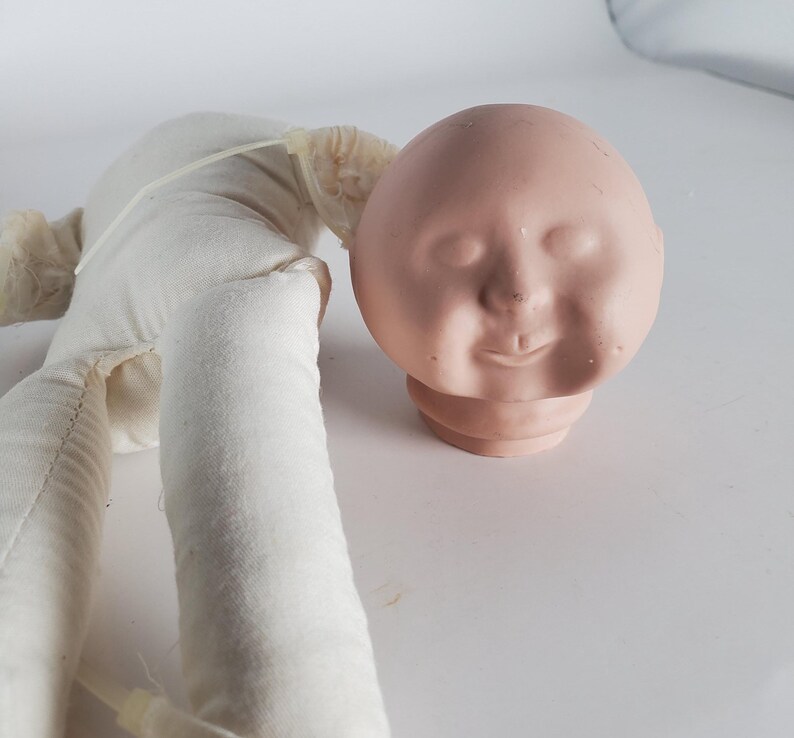 May include: A vintage doll head with a painted face and a cloth body. The doll head is a light pink color with closed eyes and a slight smile. The cloth body is white.