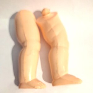 May include: Two light peach-coloured doll legs. The legs are made of a smooth plastic material. The legs are slightly bent at the knees and have detailed feet. The legs are separated and appear to be from a vintage doll.