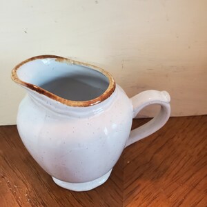 May include: A white ceramic creamer with a brown rim and speckled glaze. The creamer has a rounded body and a small handle.