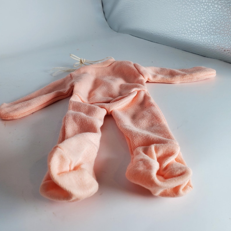 May include: A peach-colored doll outfit, featuring a long-sleeved top and pants. The outfit is made of a soft, knitted fabric. The sleeves and pant legs are extended. A string is attached at the neck.