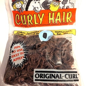May include: A clear plastic bag filled with brown curly hair strands. The bag has the text "CURLY HAIR" in orange letters. The bag also has the text "ORIGINAL-CURL" and "FUN TO USE EASY TO APPLY". The bag contains .90 oz (25 grams) of hair.