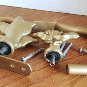 May include: Two gold metal curtain rod finials with a floral design. The finials are attached to a gold metal curtain rod. The finials are missing screws.