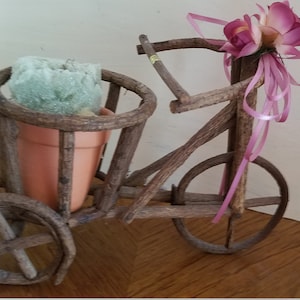 May include: A small wooden tricycle with a basket holding a terracotta pot and a green plant. The tricycle is decorated with a pink ribbon and two pink flowers.