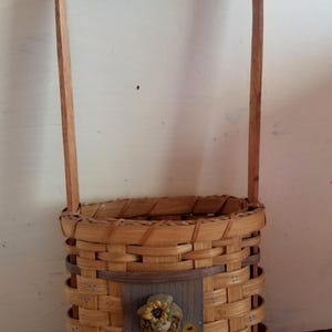 Peut inclure: Panier tressé avec une poignée en bois. Le panier présente un panneau décoratif avec une figurine d'une fille avec un chapeau de tournesol, entourée de tournesols. Le panier est fait de matériau tissé brun clair.