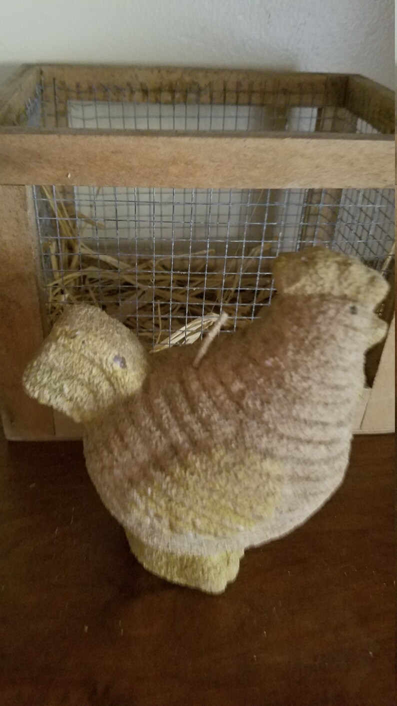 Pode incluir: Uma vela em forma de frango amarela e marrom, sentada dentro de uma gaiola de madeira com um ninho de palha.