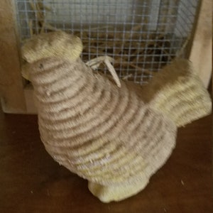 Pode incluir: Uma figurinha de frango de palha tecida com bico e p&eacute;s amarelos. O frango est&aacute; sentado em uma superf&iacute;cie de madeira.