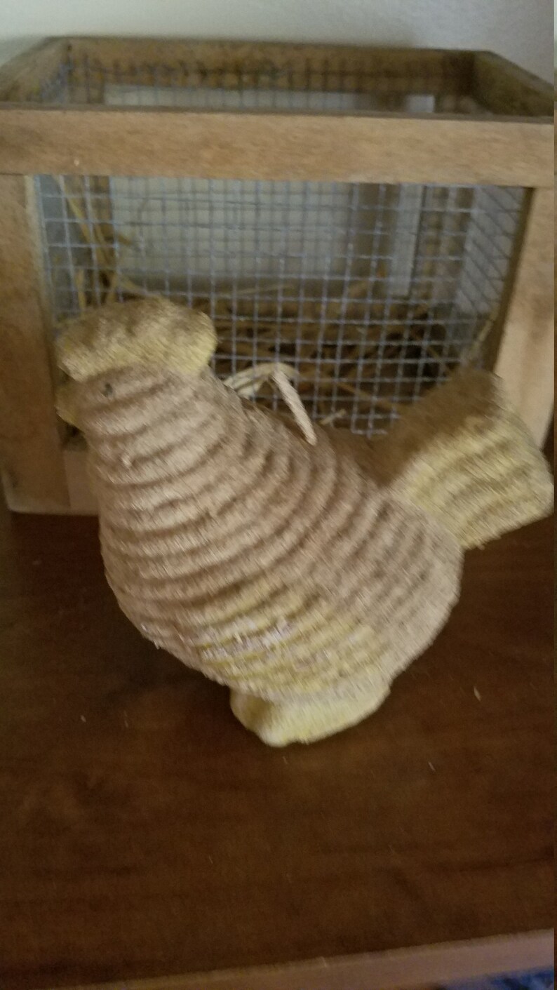 Pode incluir: Uma figurinha de frango de palha tecida com bico e p&eacute;s amarelos. O frango est&aacute; sentado em uma superf&iacute;cie de madeira.