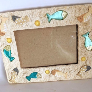 May include: A rectangular photo frame with a sandy texture, adorned with colorful fish and yellow accents. The frame features a central opening for a photo and is designed with a coastal theme.