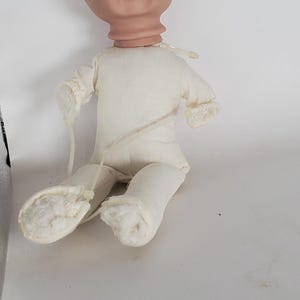 May include: A doll-making supply featuring a bisque-colored head with a smiling expression, attached to a cream-colored fabric body. The doll's limbs are also made of the same fabric. Clear plastic ties secure the limbs.