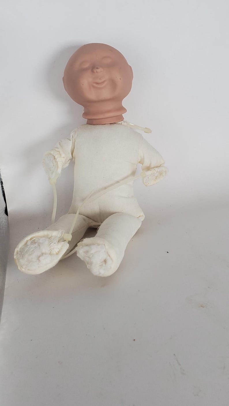 May include: A doll-making supply featuring a bisque-colored head with a smiling expression, attached to a cream-colored fabric body. The doll's limbs are also made of the same fabric. Clear plastic ties secure the limbs.