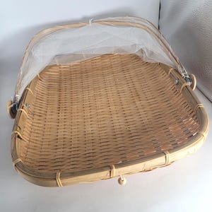 May include: A woven bamboo food cover with a white mesh net. The basket is square with rounded corners and a hinged handle. The basket is made of light brown bamboo.