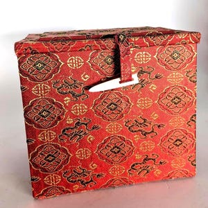 May include: A red fabric box with a repeating floral and geometric pattern in gold and black. The box has a rectangular shape with a hinged lid and a white clasp closure. The fabric has a textured appearance.