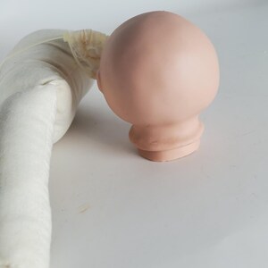 May include: A light pink doll head with a rounded shape and a detailed neck. The head is smooth and appears to be made of a solid material. A white fabric doll body is partially visible on the left side of the image.