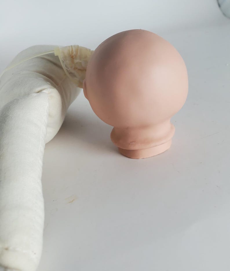 May include: A light pink doll head with a rounded shape and a detailed neck. The head is smooth and appears to be made of a solid material. A white fabric doll body is partially visible on the left side of the image.