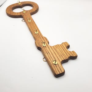 May include: A wooden key-shaped key holder with six brass hooks. The key holder is made of light brown wood with a grain pattern.
