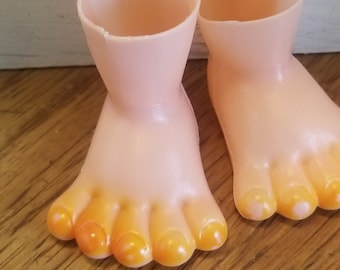 May include: A pair of plastic doll feet with orange painted toes. The feet are pink and have a slight shine.