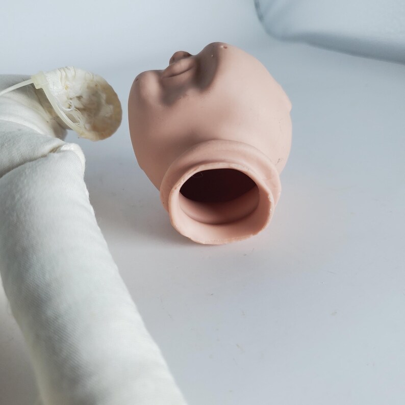May include: A doll head with a light skin tone, featuring a detailed face and open neck. The head is positioned next to a white fabric-covered body part, suggesting a craft or doll-making project.