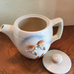 May include: White ceramic teapot with a blue and brown design featuring two birds in flight. The teapot has a lid with a knob on top.
