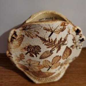 May include: A small, white ceramic vase with a brown leaf design. The vase has a unique, irregular shape and a handle on the top. The text "Dirty Sally's" is visible on the vase.