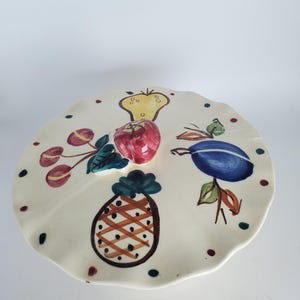 May include: A cream-colored ceramic serving plate with a scalloped edge, decorated with hand-painted fruit. The plate features a red apple, a yellow pear, a blue plum, a pineapple, and cherries. The plate is adorned with small, colorful polka dots.