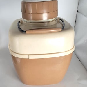 May include: A vintage, two-tone insulated cooler in beige and brown. The cooler has a rounded rectangular shape with a brown lid and base, and a beige midsection. It features a brown handle and a black carrying handle.
