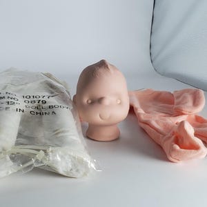 May include: A doll-making kit featuring a light pink doll head, a pair of peach-colored doll legs, and a pair of peach-colored doll legs. The doll head has a simple, rounded design. The doll legs are in a plastic bag.
