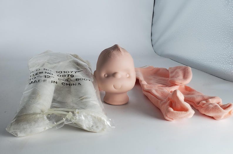 May include: A doll-making kit featuring a light pink doll head, a pair of peach-colored doll legs, and a pair of peach-colored doll legs. The doll head has a simple, rounded design. The doll legs are in a plastic bag.
