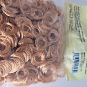 May include: A package of light brown doll hair in tight ringlet curls. The hair is made of polypropylene filament. The package has a yellow label with text and a barcode. The text on the label includes a warning to keep out of reach of children.