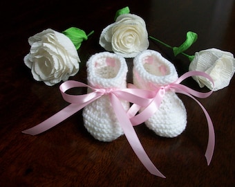 May include: Two white crocheted baby booties with pink satin ribbon tied around them. The booties are surrounded by white paper roses.