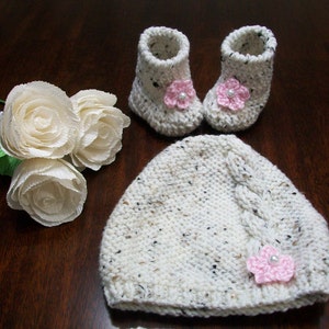 May include: A white crocheted baby hat with a pink flower embellishment, a pair of white crocheted booties with pink flower embellishments, and three white paper roses.