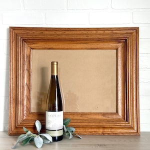 May include: A rectangular wooden picture frame with a medium brown finish and a prominent wood grain texture. In front of the frame stands a tall, clear glass wine bottle with a white label reading "La Chapelle" and a small sprig of green leaves.