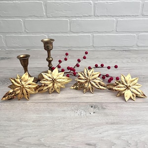 May include: A festive holiday centerpiece featuring gold poinsettia flowers, red berries, and brass candle holders. The arrangement is set against a light gray wooden surface and a white brick wall, creating a warm and inviting atmosphere.