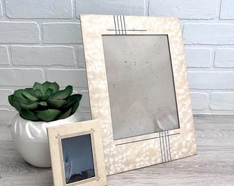 May include: Two vintage picture frames. The larger frame has a light beige speckled pattern with black vertical lines. The smaller frame is rectangular with a similar design. A green succulent plant in a white pot is also in the image.