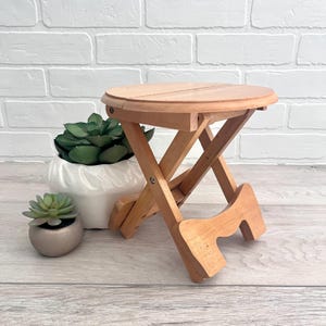 May include: A small, foldable wooden stool with a round top. The stool is made of light-colored wood and has an X-shaped base. The stool is approximately 12 inches tall and is suitable for indoor or outdoor use.