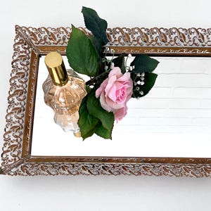 May include: A gold-colored metal tray with a mirrored surface and an ornate design. A clear glass bottle of perfume with a gold cap and a pink rose are on the tray.