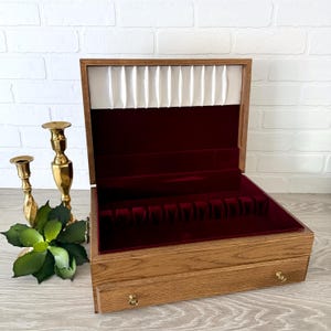 May include: Open wooden silverware chest with a red velvet interior and a white satin lining. The chest has a light brown finish and brass hardware. Two gold candlesticks and a green plant are next to the chest.