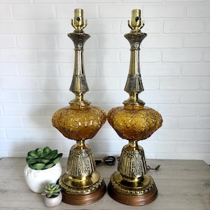May include: A pair of matching table lamps featuring amber-colored glass bodies with intricate embossed floral patterns. The lamps have polished brass metal stems and bases, topped with standard light bulb sockets, standing on circular wooden bases.