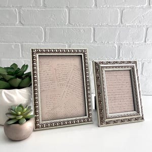 May include: Two silver picture frames with ornate designs. The larger frame is on the left and the smaller frame is on the right. Both frames contain handwritten letters on a light pink background.